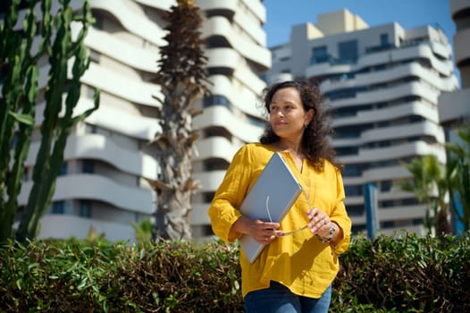 Confident portrait of an ethnic businesswoman, copywriter with laptop, looking away against modern buildings background
