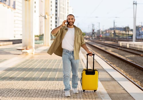 Positive millennial european guy with suitcase calls by phone, talk, waiting for train