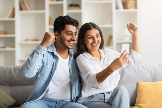Excited indian couple celebrating, looking at smartphone