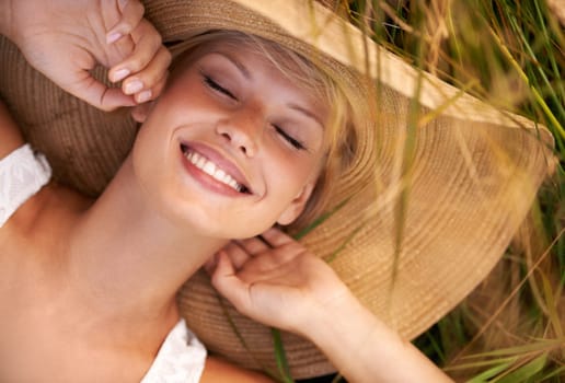 Woman, hat and relax on grass with peace, happiness and freedom in summer. Outdoor, fashion and girl lying on lawn with a smile on face for holiday, vacation or free time in nature with carefree joy