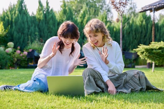 Teenage female student friends laughing sitting on grass with laptop