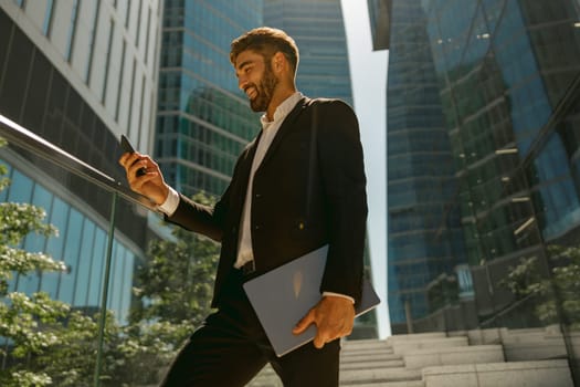 Handsome businessman using mobile phone standing with laptop on background of skyscrapers
