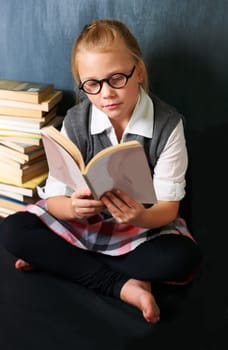 Glasses, reading and child student with a book in the classroom for education, learning or knowledge. Studying, nerd and girl kid enjoying a story, fantasy or novel in library at school or academy.