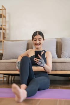 Lifestyle asian young fitness woman holding smartphone relaxing after workout at home. Smiling female using cell phone checking newsfeed on social media while exercise