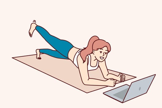 Woman is doing fitness lying on yoga mat and looking at laptop, listening to advice online trainer