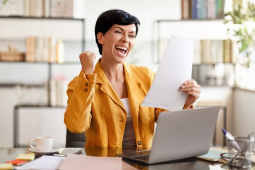 European businesswoman shaking fists at laptop holding papers at office