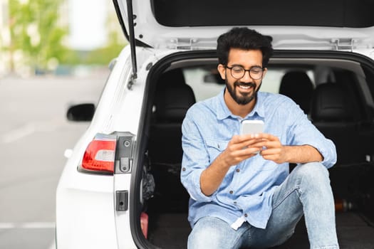 Happy indian guy using cell phone, sitting on car trunk