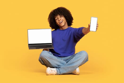 Cheerful Black Guy Showing Laptop And Smartphone With Blank White Screen