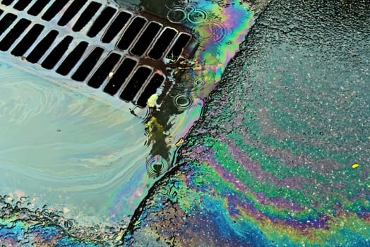 An oil slick against the backdrop of an asphalt road flows into a storm drain through a grate
