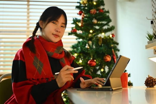 Pretty young woman in warm clothes using digital tablet sitting by decorated Christmas tree in living room.
