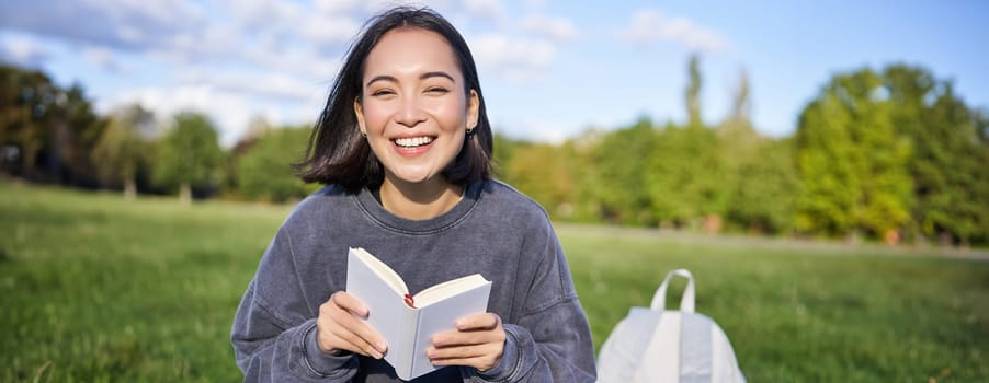 Beautiful asian girl sitting in park on grass, reading and smiling. Woman with book enjoying sunny day outdoors.