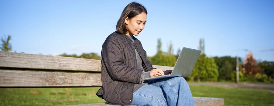 Digital nomad. Portrait of young woman using laptop in park, sitting on bench and working, studying online.