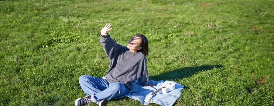 Happy asian girl sitting on picnic blanket with ukulele, cover herself from sunlight, extending hand towards sun beams and smiling.