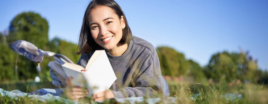 Portrait of cute korean girl, reading in park while lying on grass, relaxing with favorite book in hands, smiling happily.