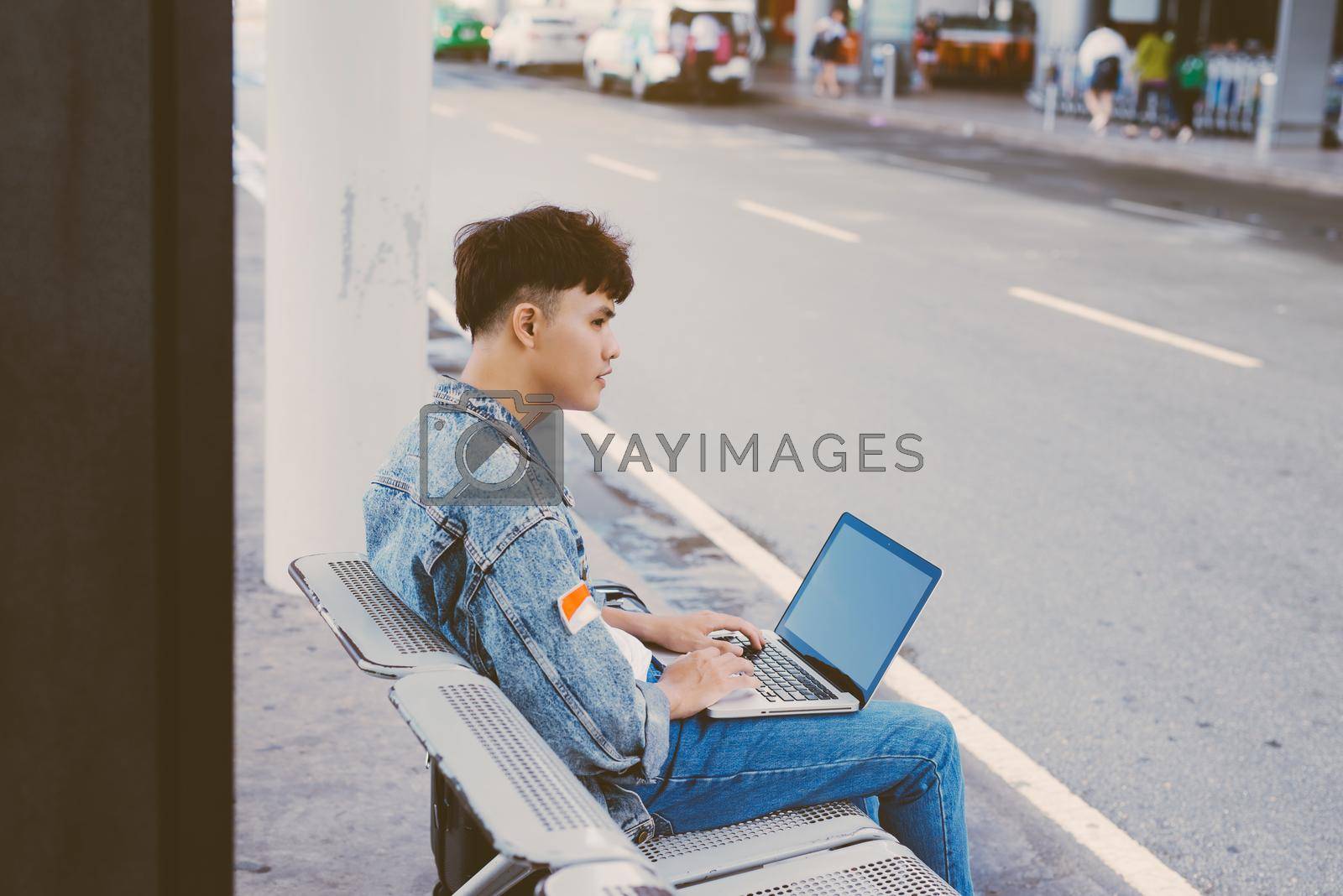 Royalty free image of Asian young man sitting on the chair at the airport bus stop and lusing laptop, side view by makidotvn