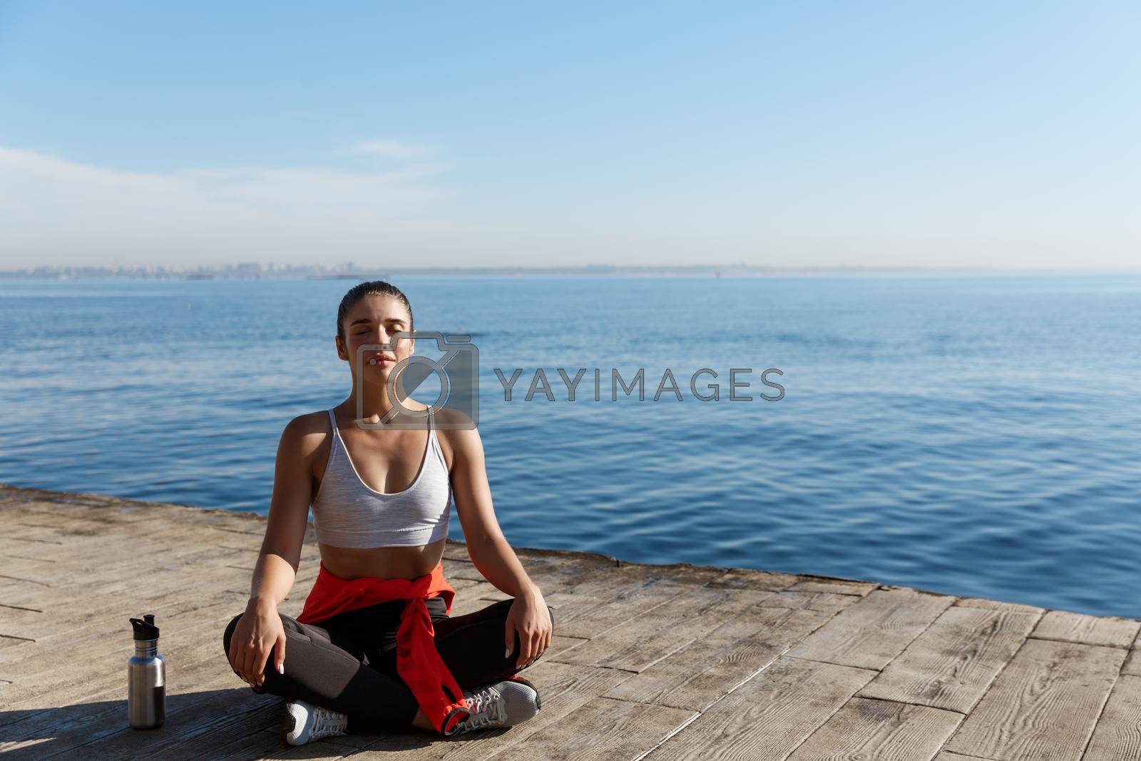 Outdoor shot of peaceful fitness woman sitting in asana and doing yoga near the sea. Female athlete meditating on seaside.