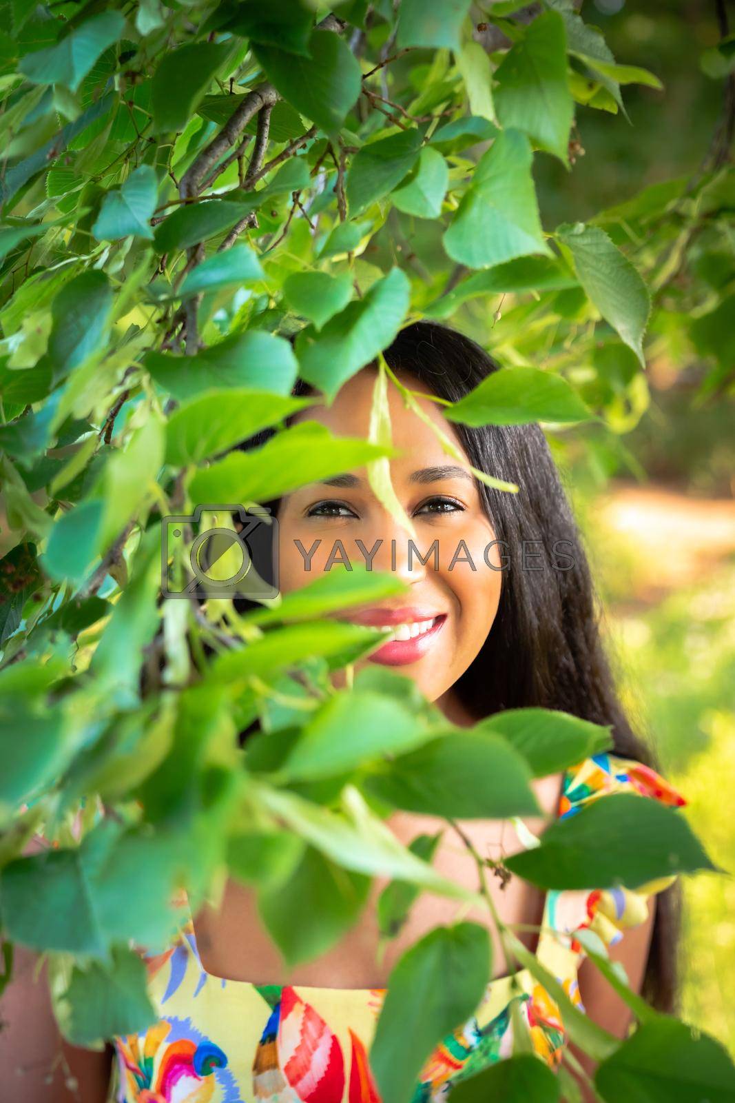Royalty free image of A beautiful young mixed race African American woman smiles with her teeth from behind a tree branch with leaves obscuring her face and upper body as she wears a yellow floral print dress. by lapse_life
