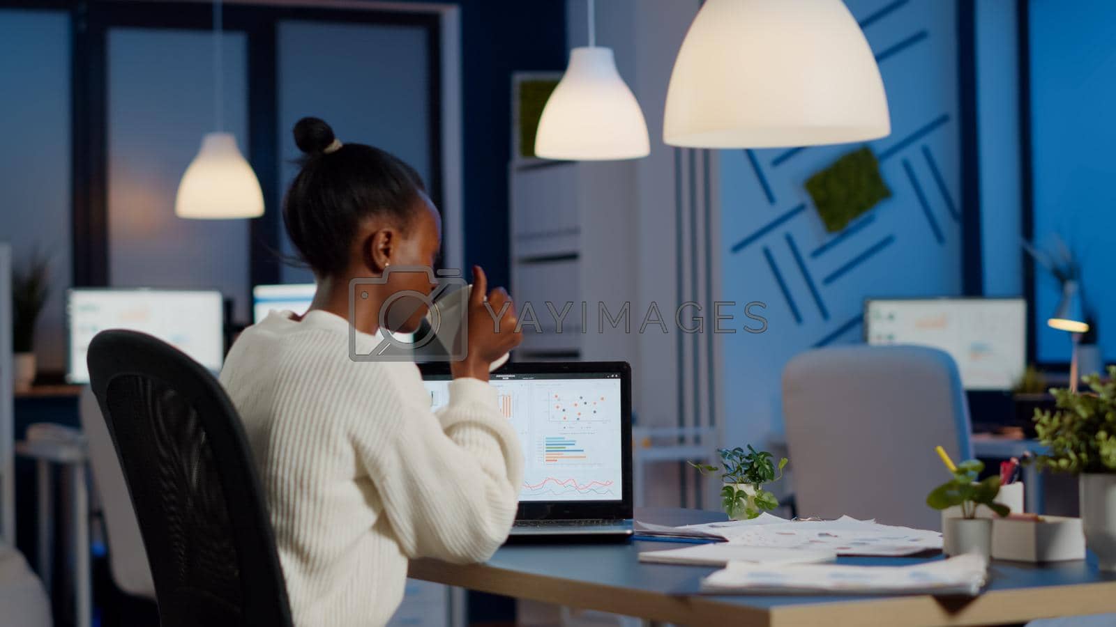 Busy african business woman analysing financial reports checking company statistics graphs, looking at laptop, pointing to numbers late at night in start-up office doing overtime to respect deadline