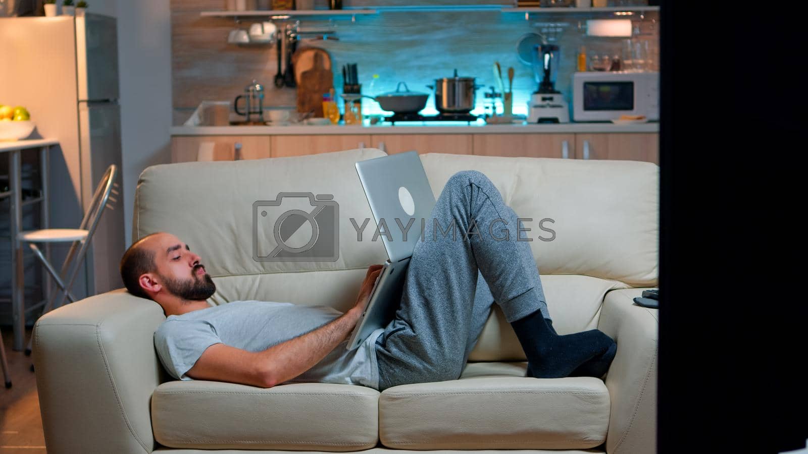 Exhausted man falling asleep while working on internet communication project using laptop computer with modern technology wireless. Caucasian male in pajamas sitting on sofa late at night in kitchen