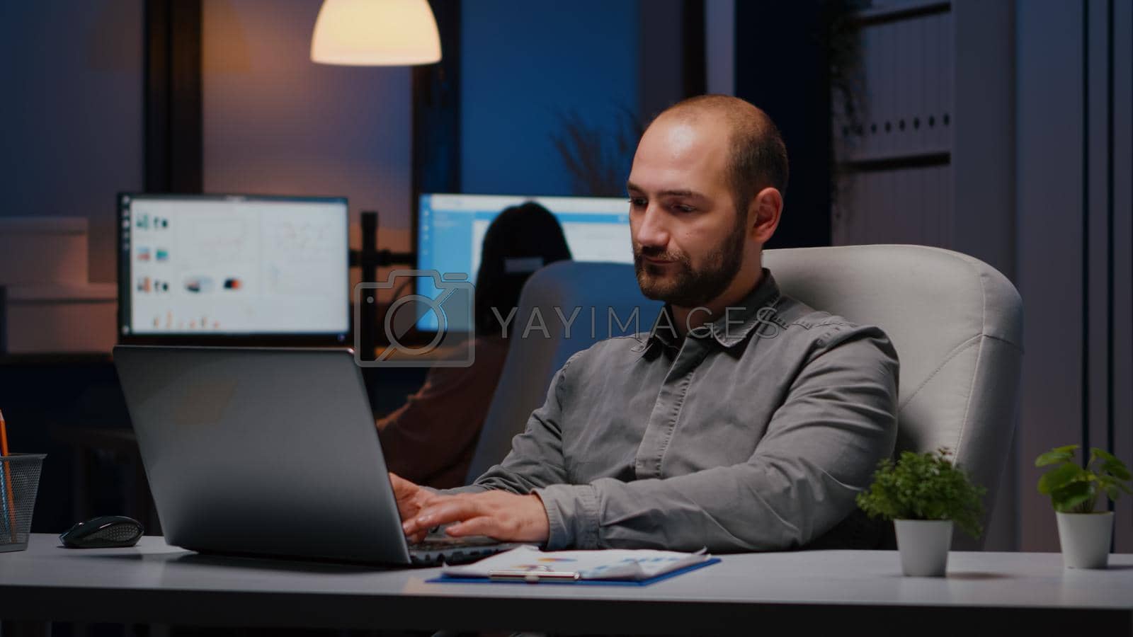 Portrait of businessman working on laptop computer planning financial strategy late at night in business company office. Workaholic manager smiling into camera while sitting on chiar