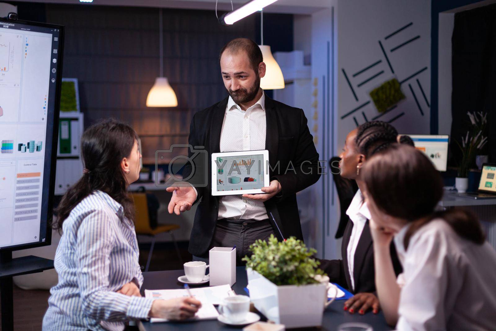 Overworked businessman showing financial graphs presentation using tablet brainstorming company ideas. Workaholic diverse multi-ethnic businesspeople overworking late at night in office meeting room