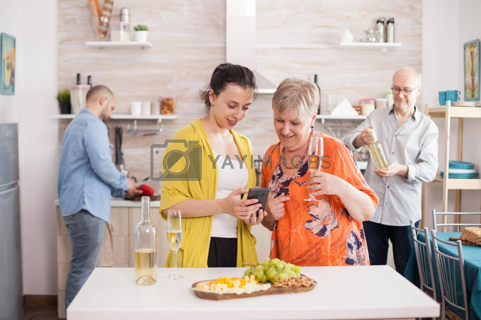 Mother and daughter browsing on smartphone in kitchen during family brunch. Senior woman holding glass of white wine. Tasty appetizer.