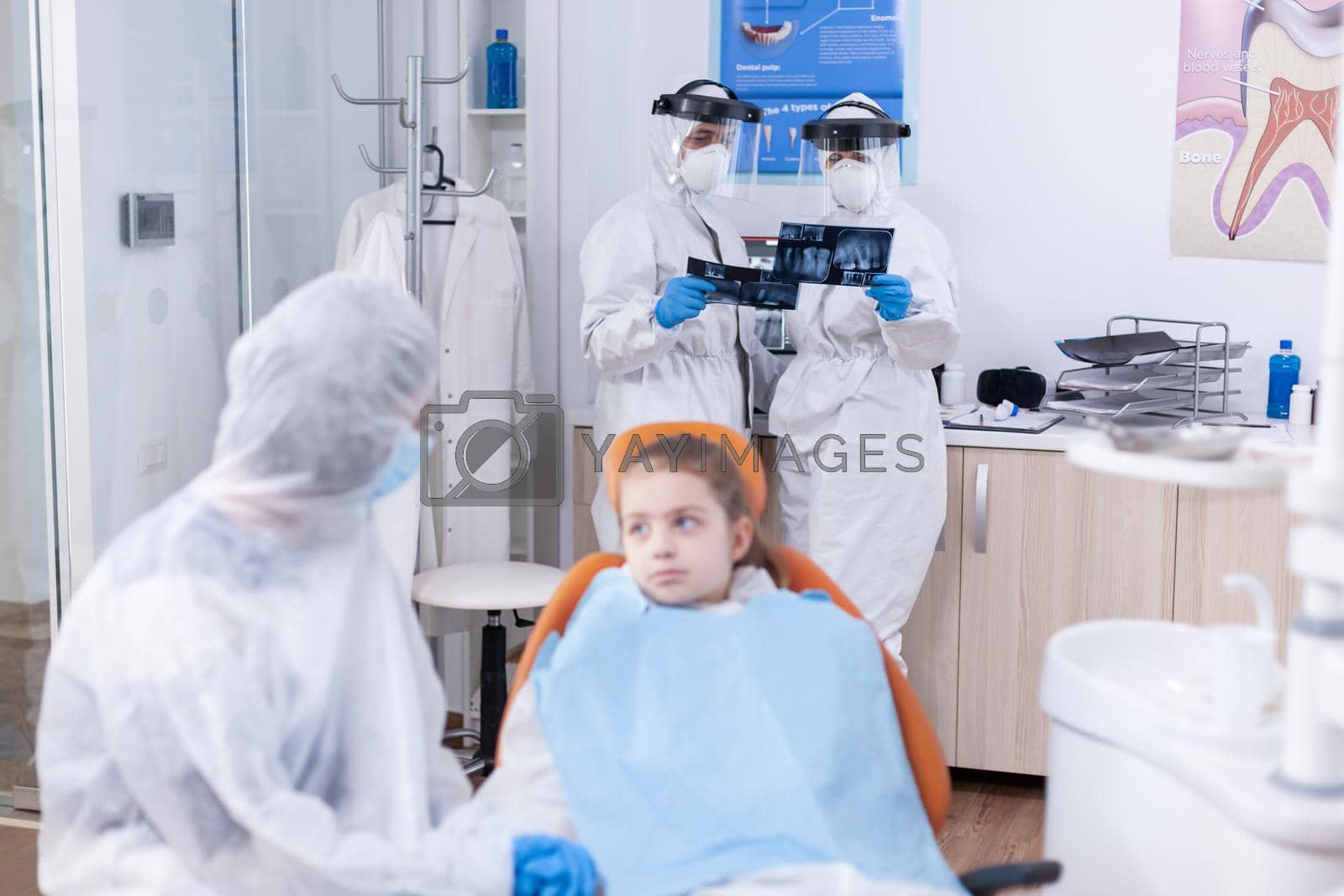 Team of dentist looking at patient dental radiography dressed in coverall in the course of coronavirus outbreak. Stomatologist during covid19 wearing ppe suit doing teeth procedure of child sitting on chair.