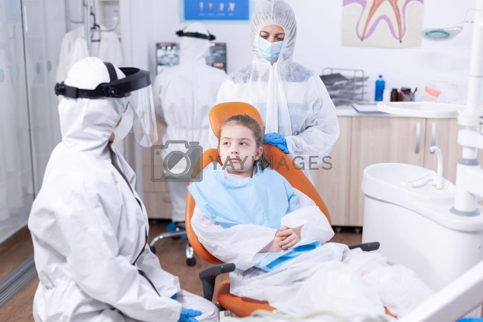 Serious little girl sitting in dental chair listening dentist talking about treatment for cavity. Stomatologist during covid19 wearing ppe suit doing teeth procedure of child sitting on chair.