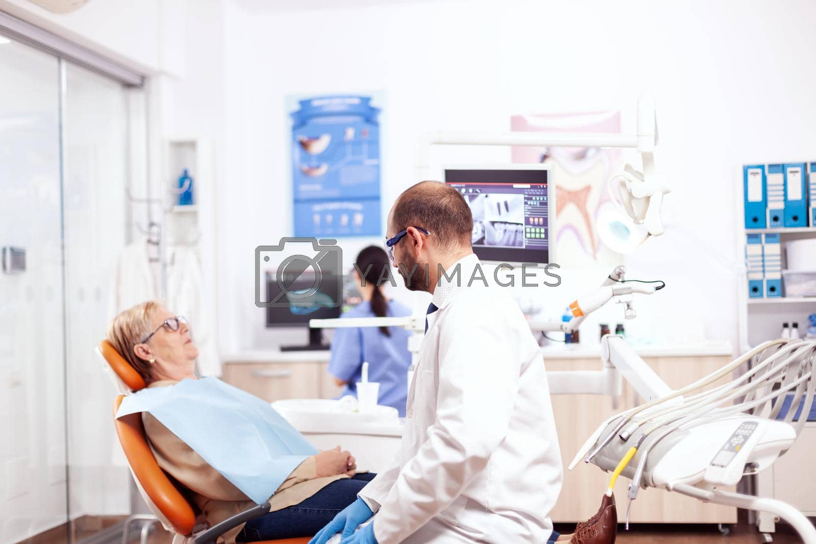 Dentist in dental clinic saying diagnostic to senior woman wearing lab coat. Medical teeth care taker talking with patient sitting on chair during consultation.