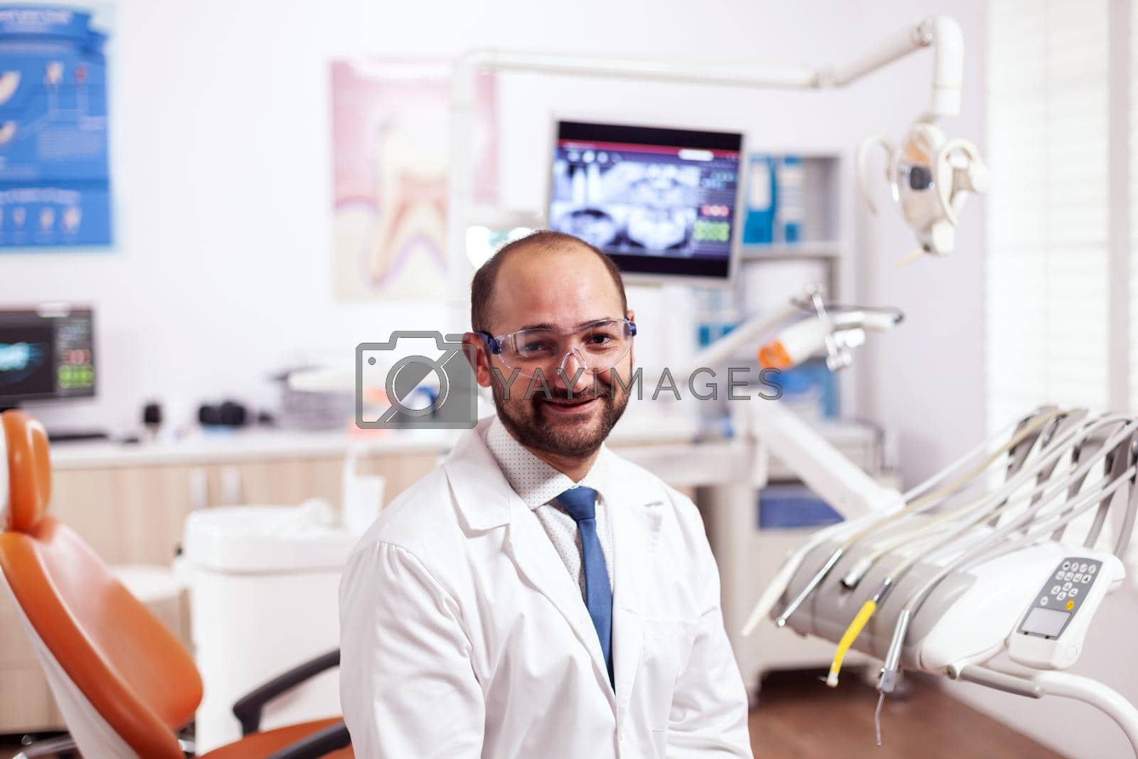 Confident dentist in stomatology cabinet with orange equiptment wearing dental uniform. Medical specialist in oral hygiene wearing lab coat looking at camera in dentistry office.