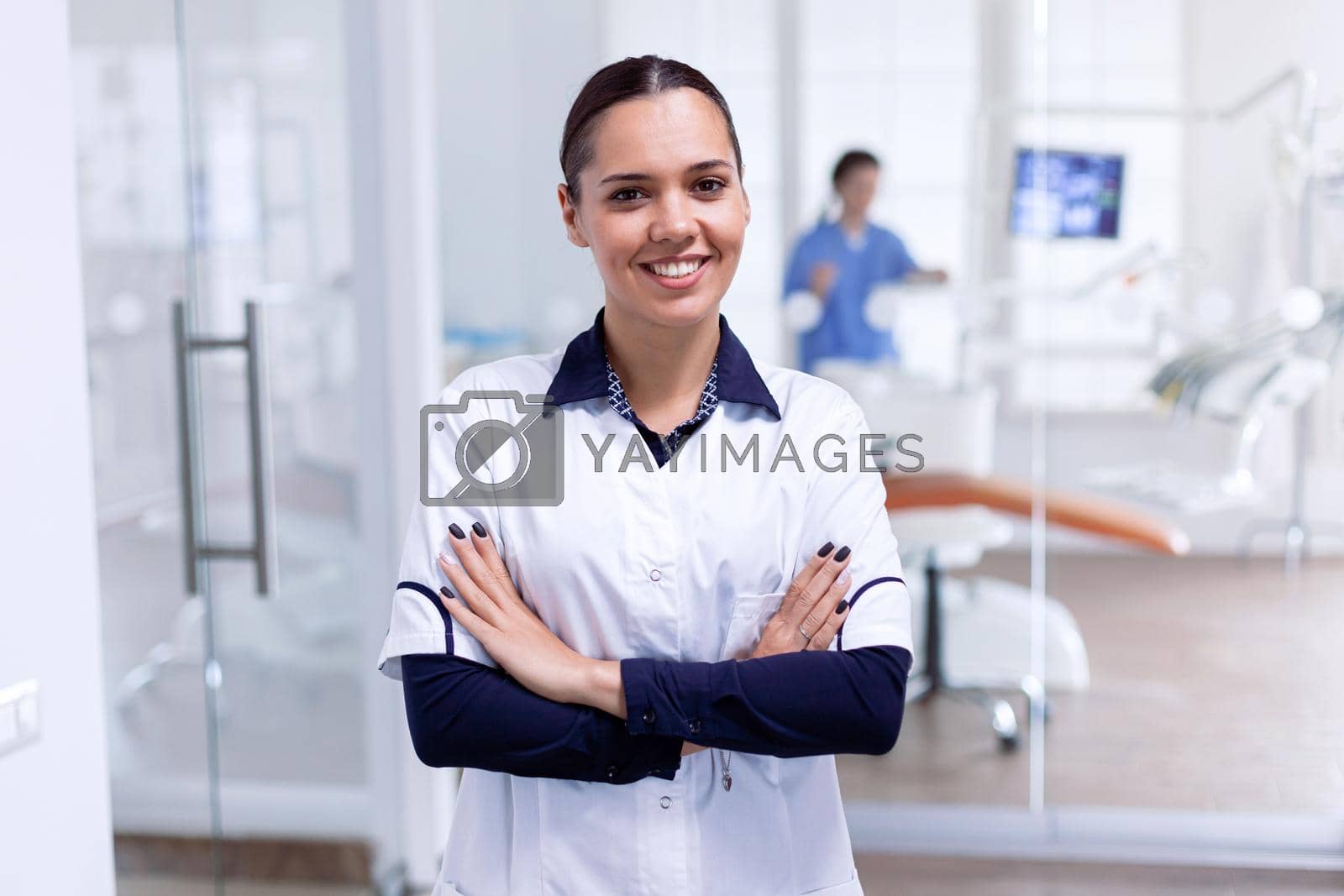 Portrait of cheerful dentist in dental reception with arms crossed looking at camera wearing uniform. Happy smiling dentist in her orthodontic office.