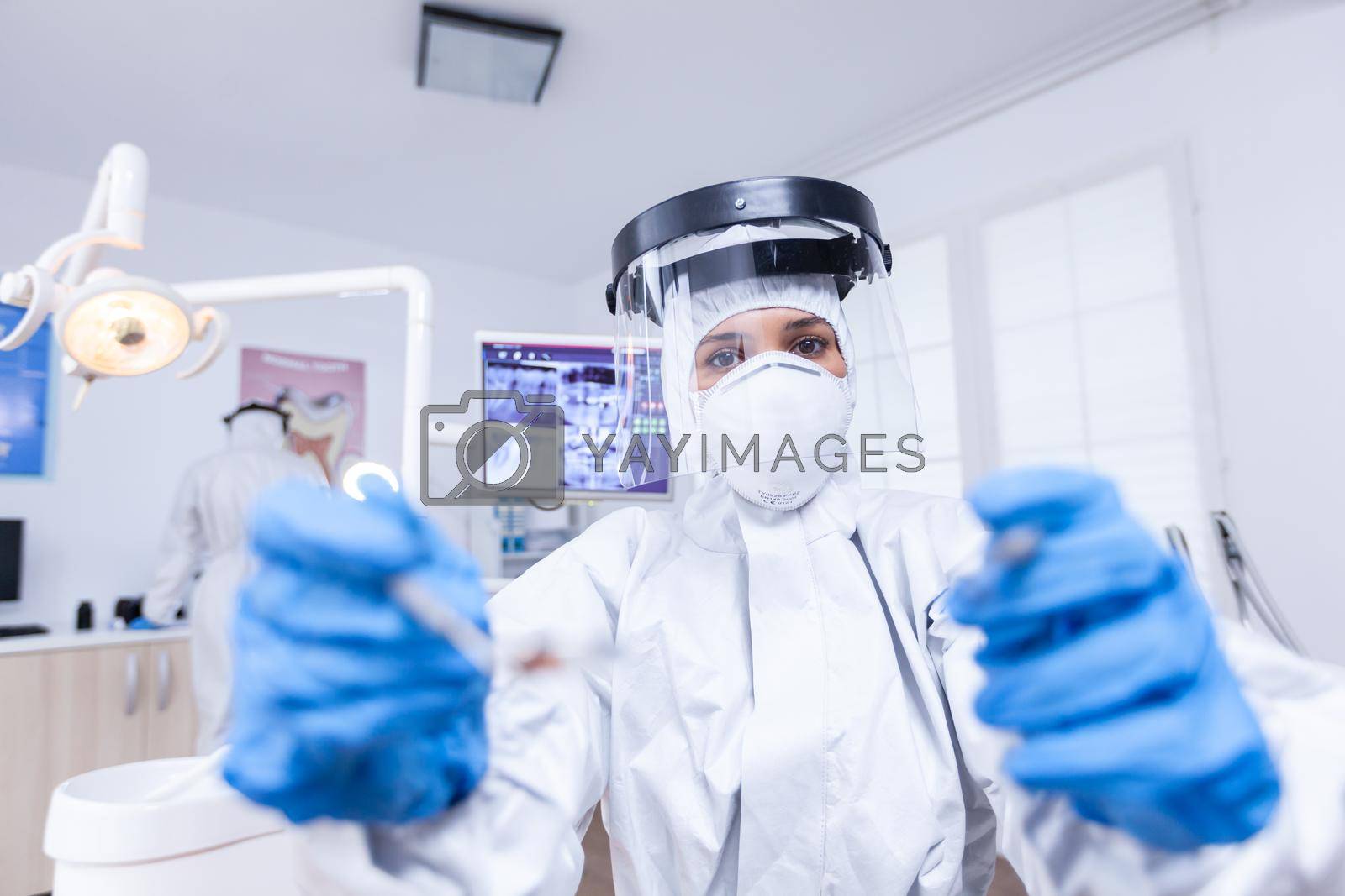 Patient pov of dentist examining patient gums using tools dressed up in covid suit in dental office. Stomatolog wearing safety gear against coronavirus during heatlhcare check of patient.