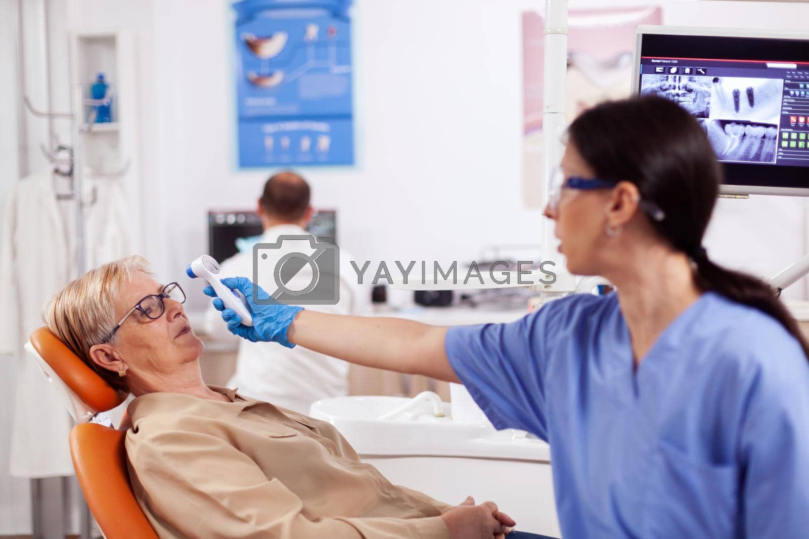 Dentist assistant measuring senior woman body temperature using thermometer during consultation. Medical specialist in dental clinic taking patient temperature using digital device.