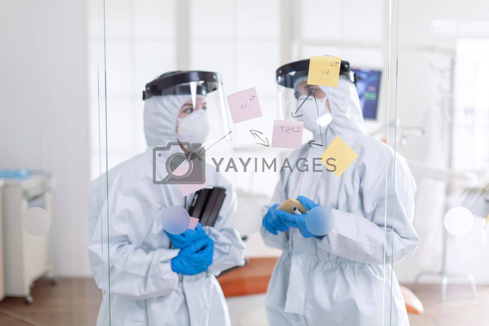 Dentist coworkers talking about teeth issues wearing pee suit as safety precauiton for covid-19. Medical team in stomatology office wearing coverall in dental office writing ideas on sticky notes.