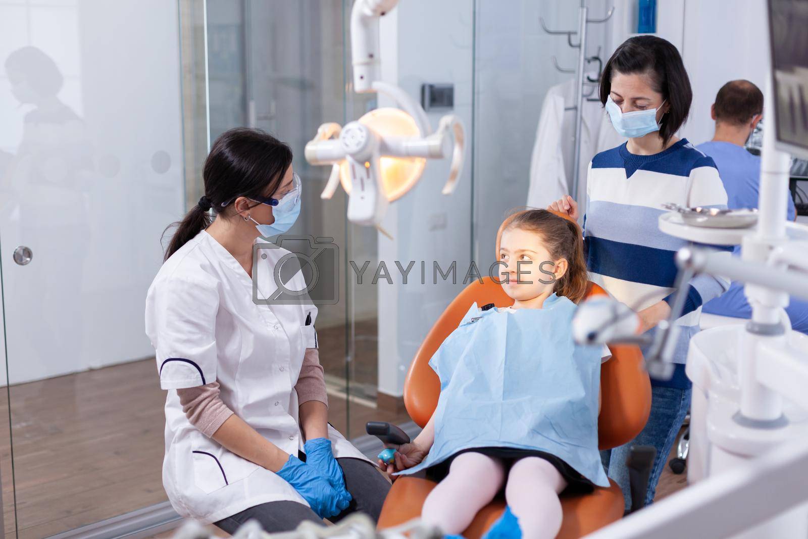 Dentist wearing face mask in the course kid examination with dental bib in dentistiry office. Dentistry specialist during child cavity consultation in stomatology office using modern technology.