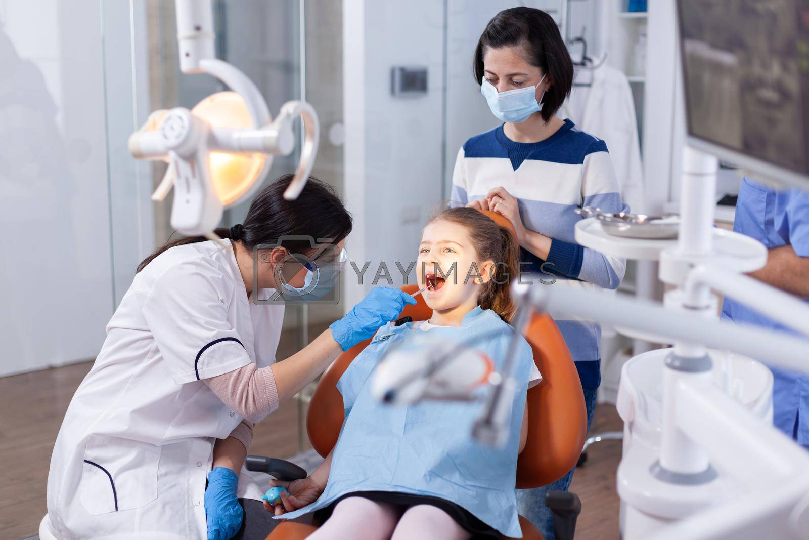 Angled mirror used by dentist doctor on little girl with mouth open in dental office. Dentistry specialist during child cavity consultation in stomatology office using modern technology.