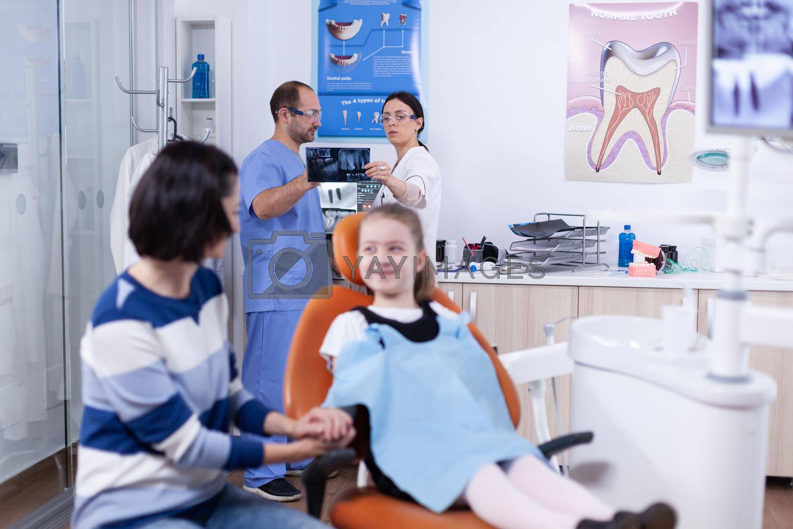 Dentist and assistant in dental office holding little girl jaw radiograph discussing diagnosis.. Child with her mother during teeth check up with stomatolog sitting on chair.