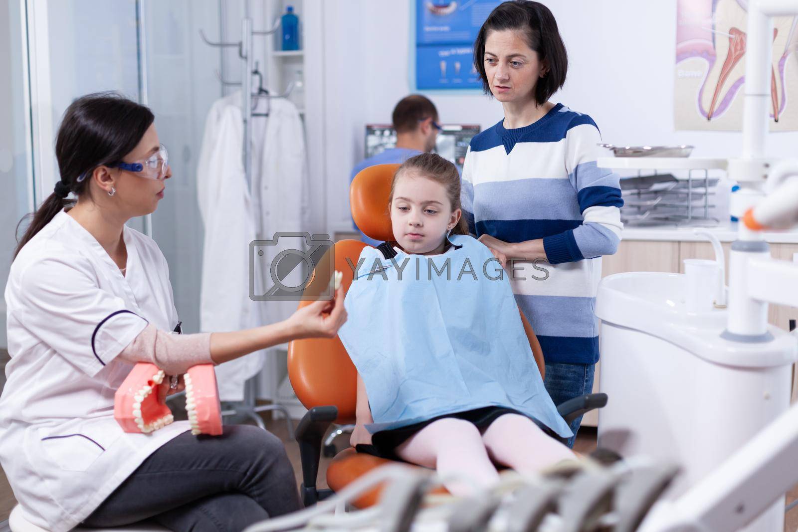 Child wearing dental bib sitting on chair while doctor is showing artificial teeth. Little girl and mother listening stomatolog talking about tooth hygine in dentistiry clinic holding jaw model.
