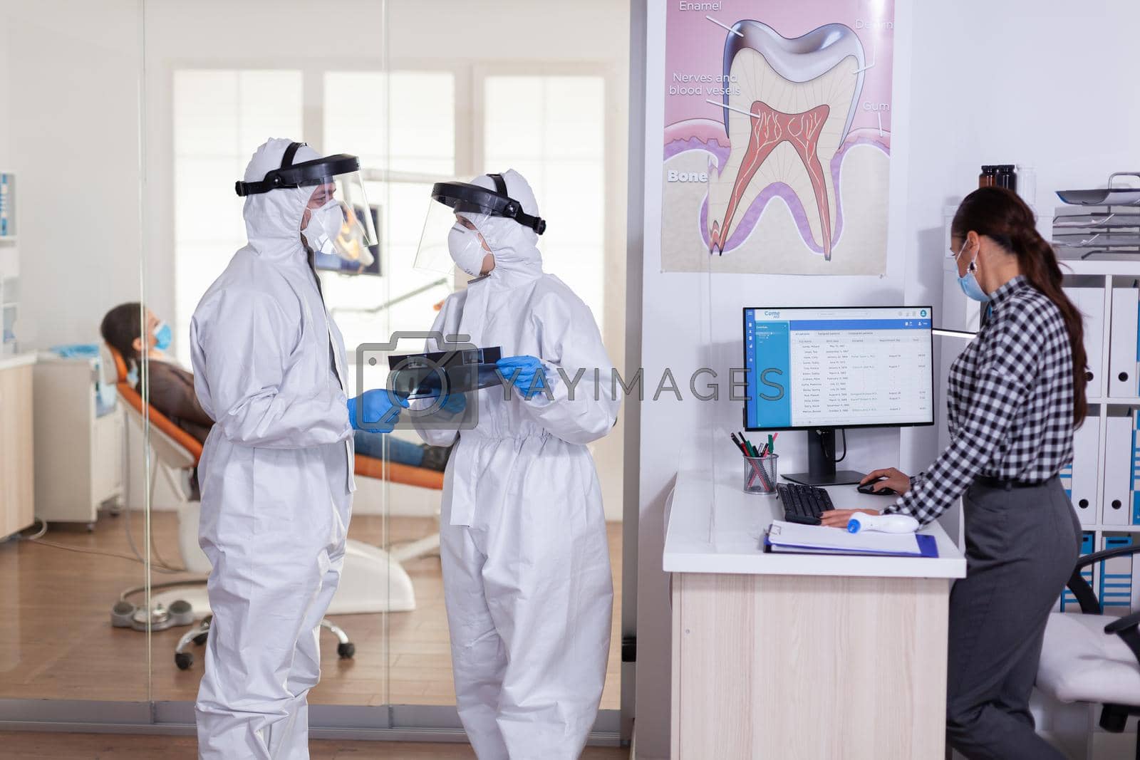 Dentist assistant discussing with doctor patient diagnosis keeping social distancing dressed in ppe suit face shiled, during global pandemic with coronavirus holding x-ray.