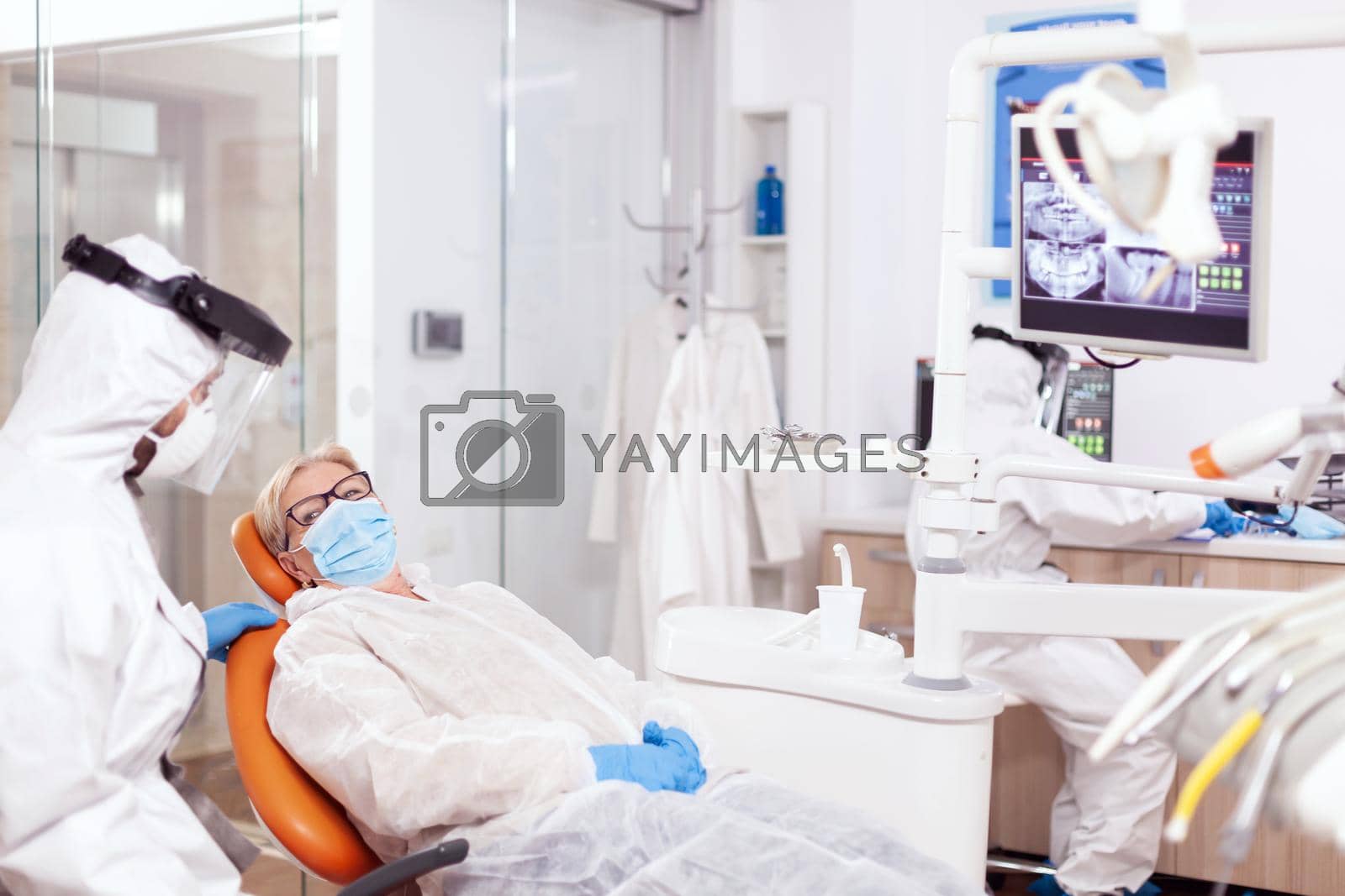 Dentist wearing protection agasint coronavirus during stomatology consultation of senior patient. Elderly woman in protective uniform during medical examination in dental clinic.