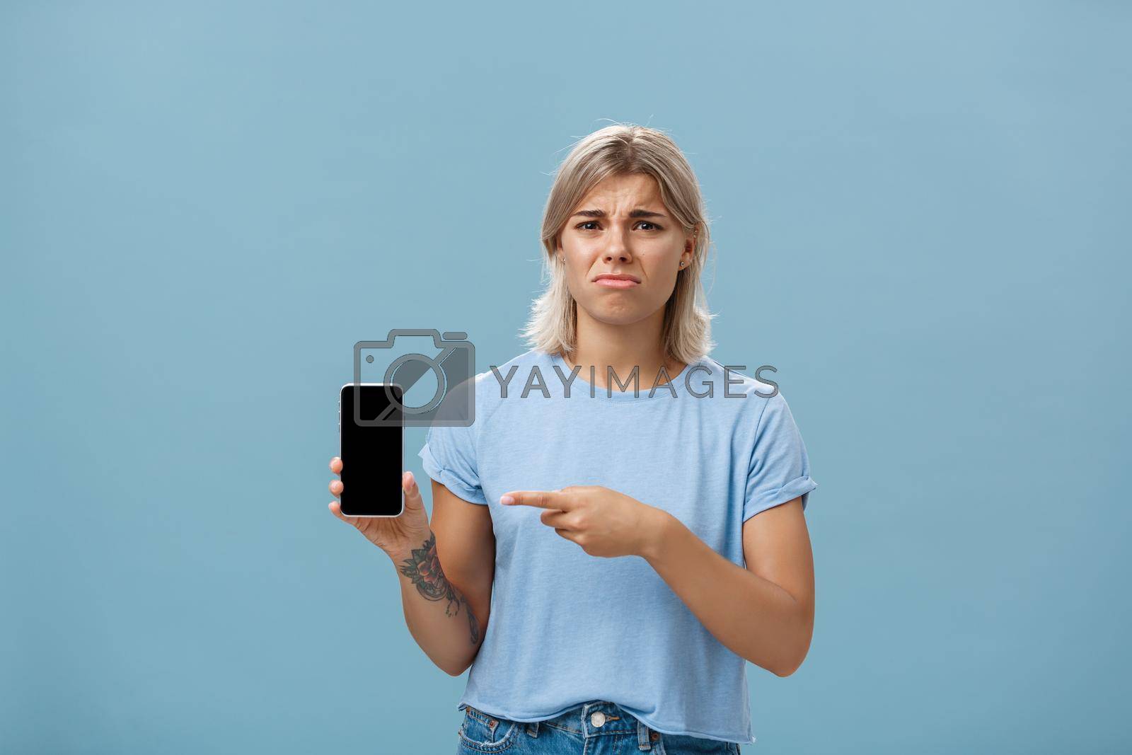 Royalty free image of Unhappy sad attractive silly girl with fair hair tattoos and tanned skin frowning with regret and disappointment poonting at smartphone screen as if being dissatisfied with new girlfriend of ex-lover by Benzoix
