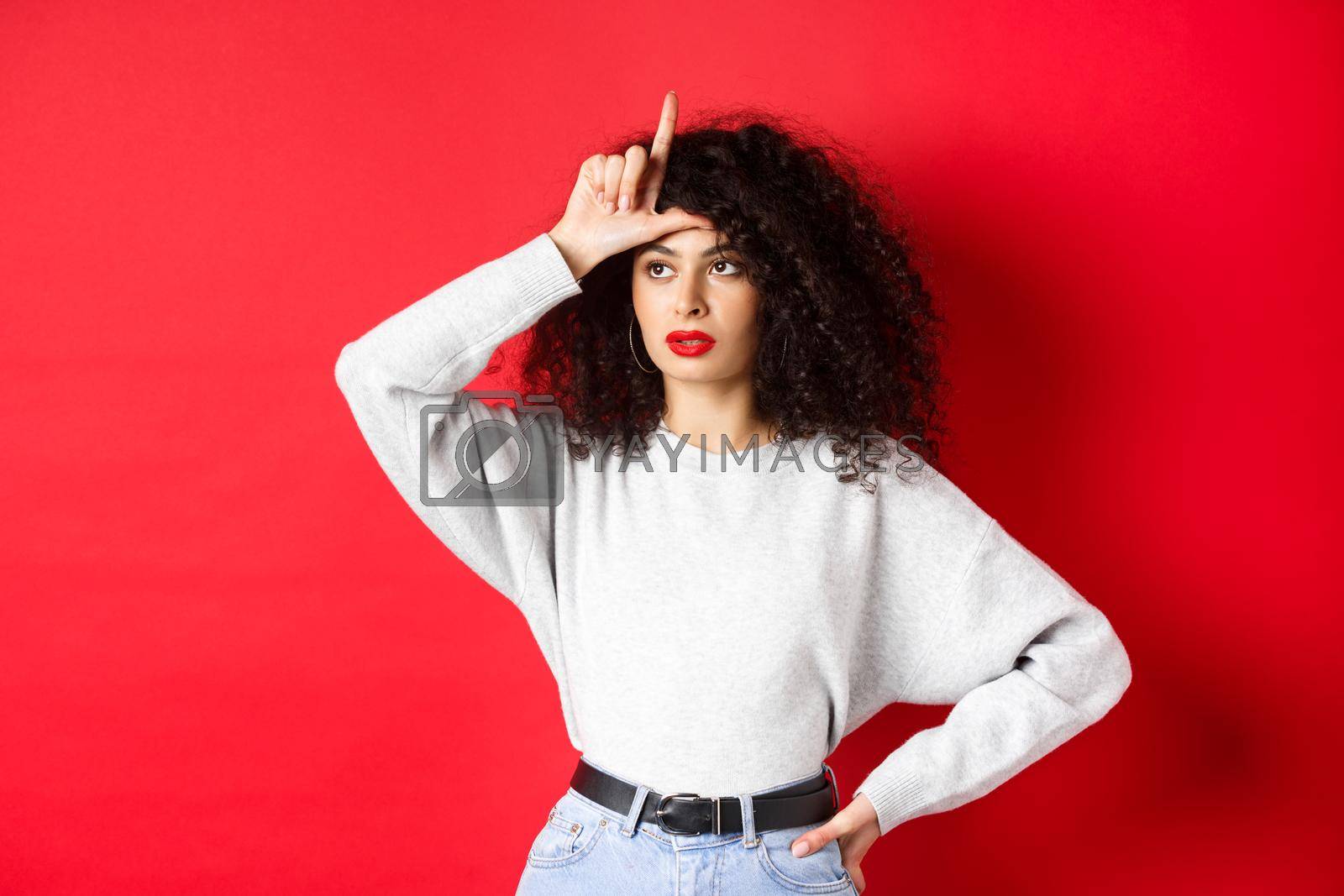 Royalty free image of Arrogant woman with curly hair, showing loser sign on forehead and looking aside, mocking someone, standing on red background by Benzoix