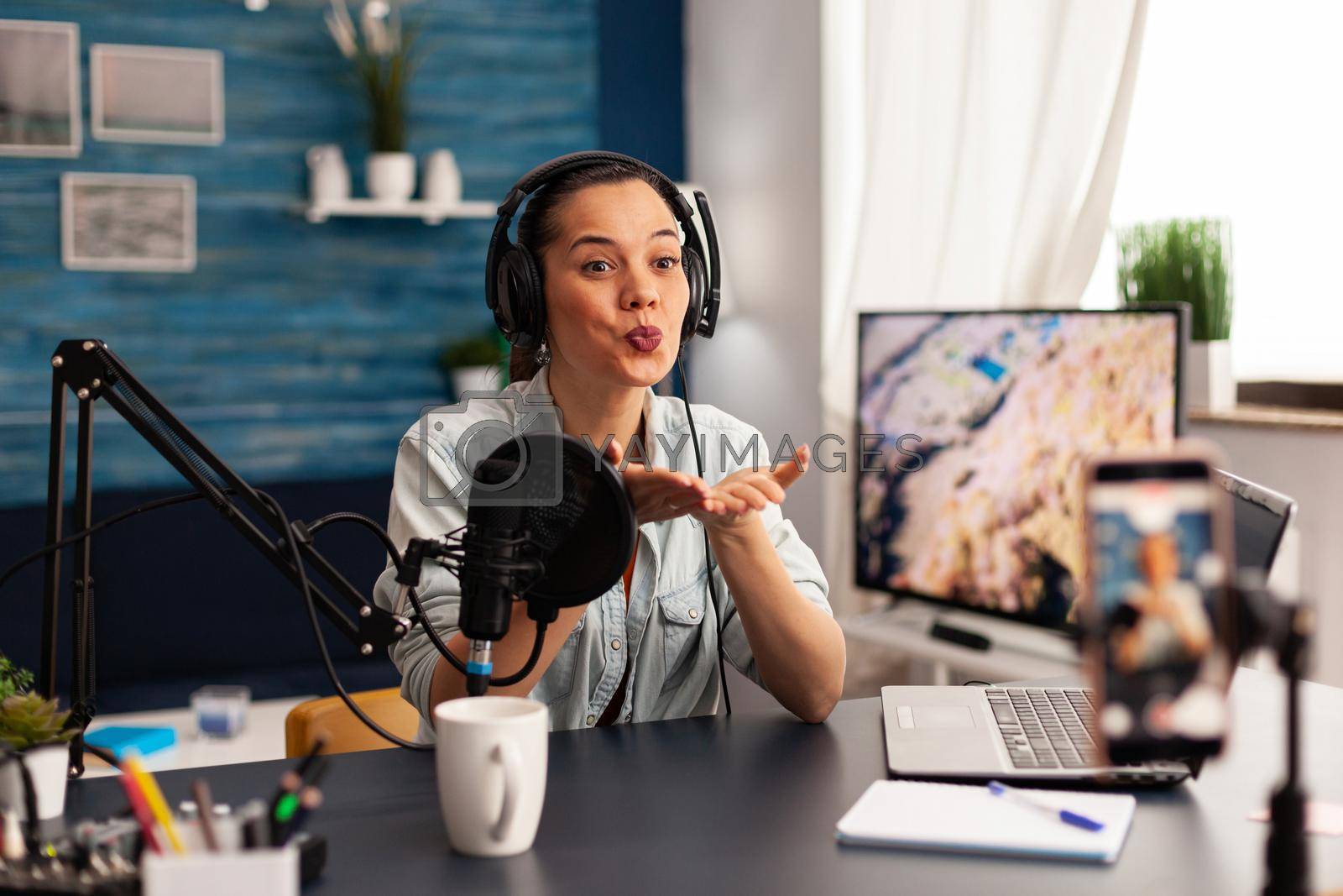 Influencer giving flying kiss while making new fashion series. Creative vlogger recording video blog concept speaking and looking at smartphone on tripod home studio podcast