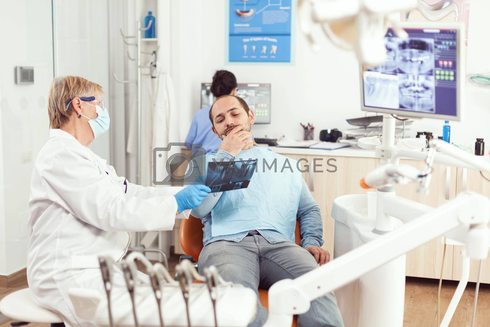 Senior woman dentist examining tooth radiography with sick man discussing toothache during stomatology appointment. Patient sitting on dental chair in hospital dentistry office