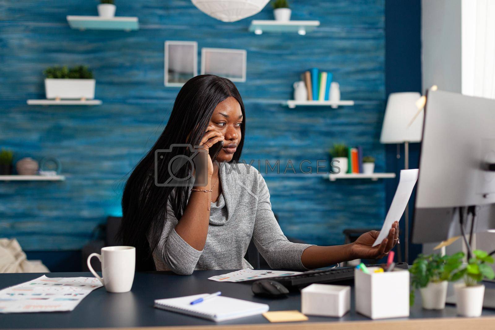 Black sad student interacting with collegue explaining communication course ideas using phone sitting at desk in living room. African woman working at academic homework using elearning platform
