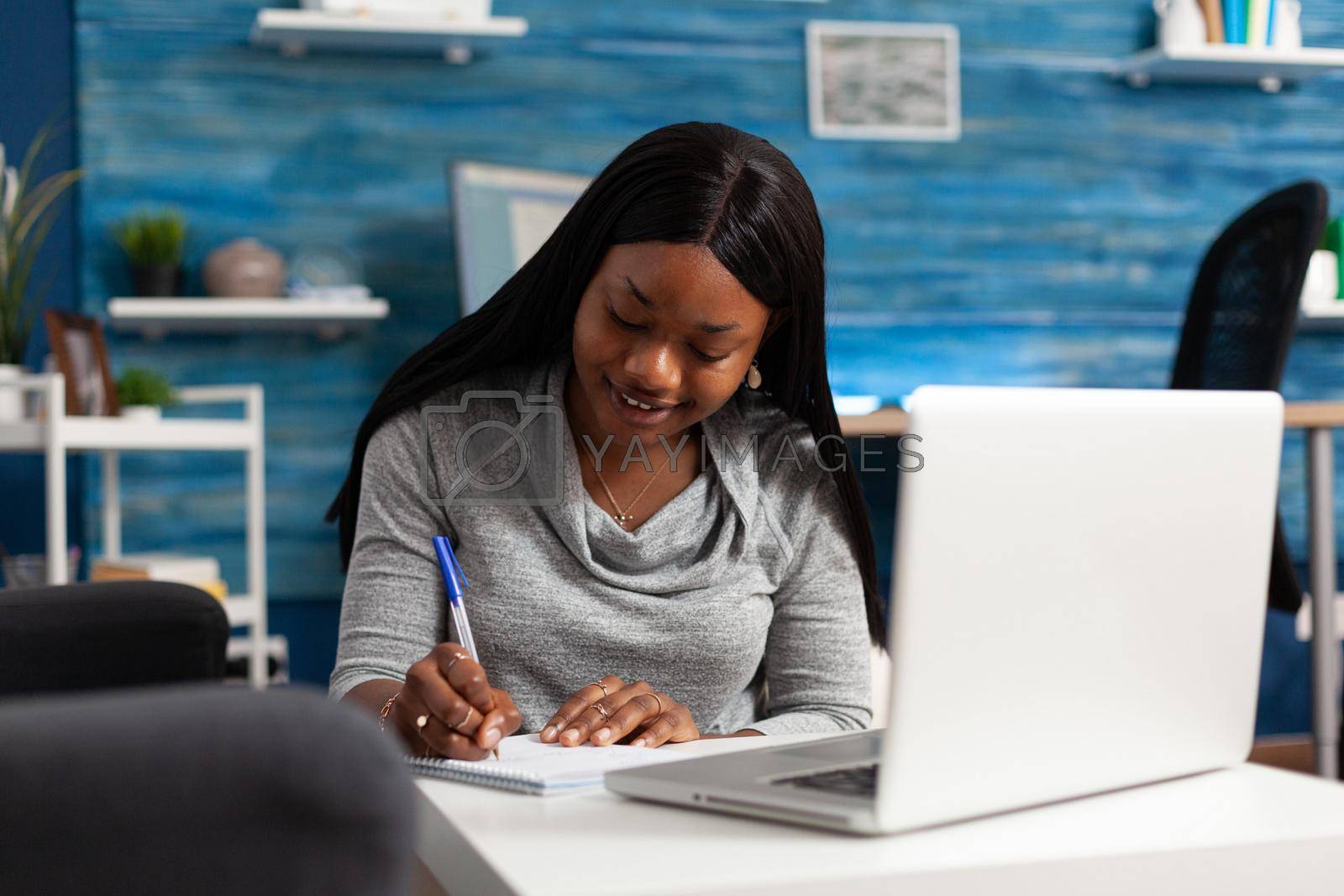 Afro american woman studying communication course writing high school lesson on notebook sitting at table in living room. Black student working remote at academic homework during university class