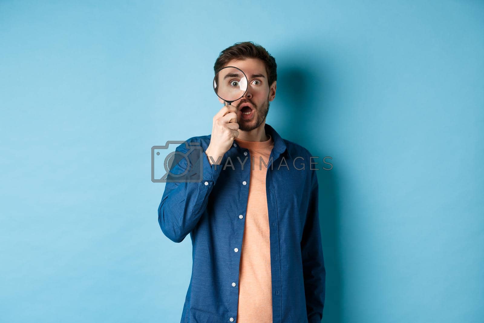 Royalty free image of Impressed man look in awe through magnifying glass, drop jaw and stare at camera, standing on blue background by Benzoix
