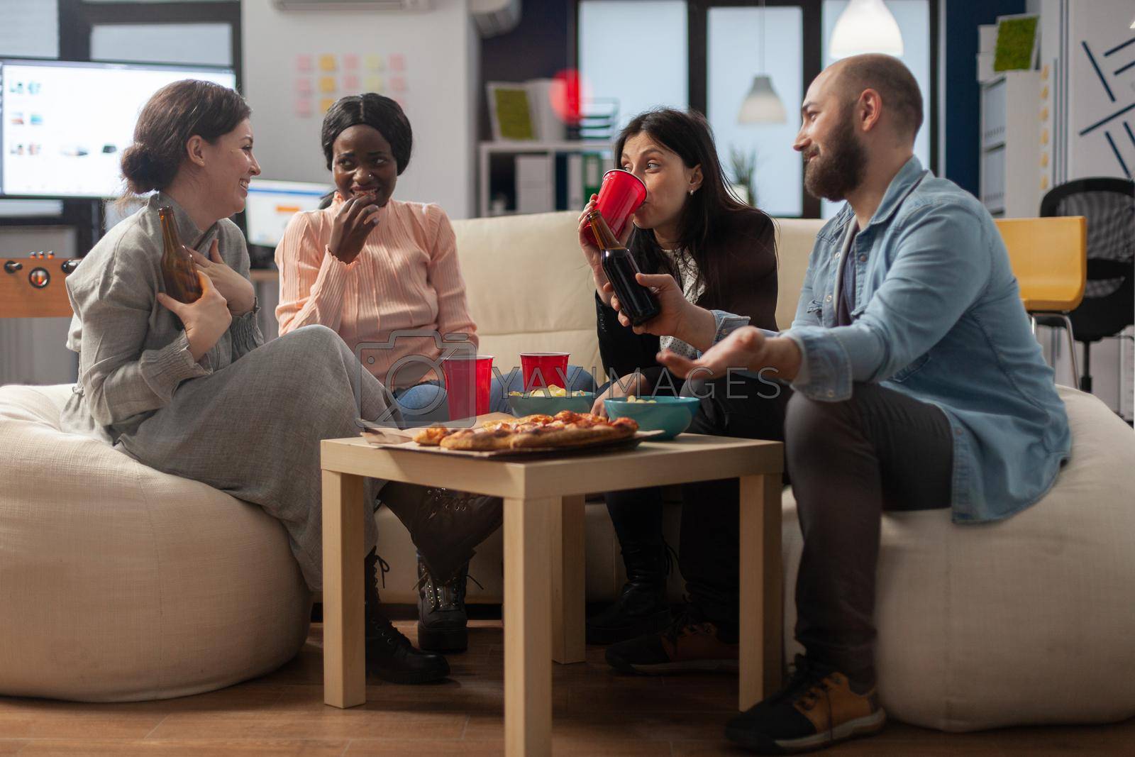 Diverse group of colleagues enjoy after work celebration drinks at office. Multi ethnic cheerful friends sitting on couch with snacks pizza chips and cups bottles of beer alcohol on table.