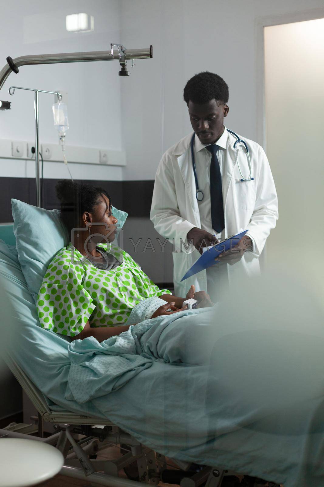 Man with doctor occupation analyzing patient healthcare in hospital ward at clinic. African american specialist, medic explaining disease, illness, diagnosis to young woman sitting in bed