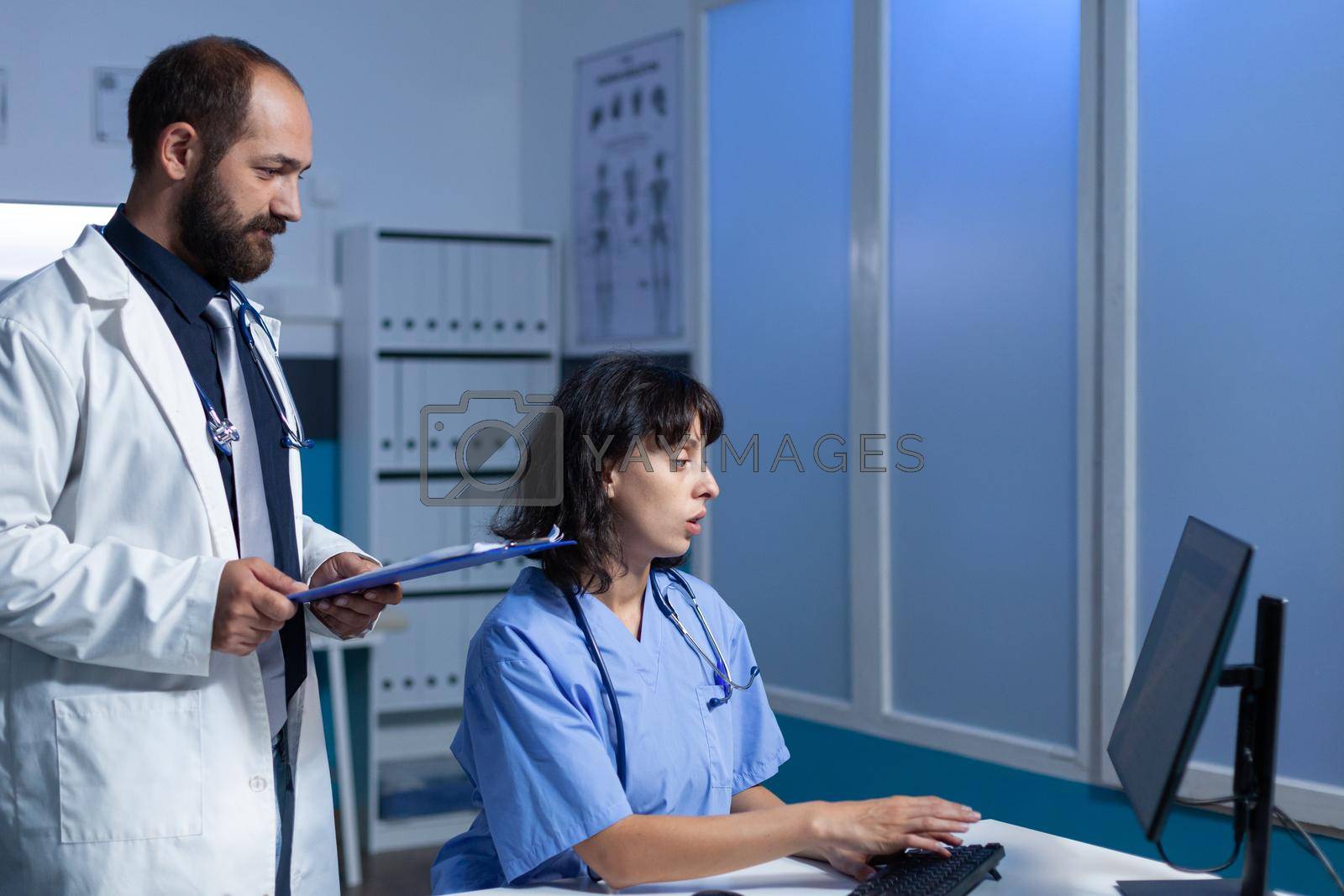 Medical team of workers looking at monitor for healthcare information and examination. Doctor holding checkup files while nurse working with computer on desk. Man and woman at job
